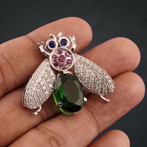 May include: A silver fly brooch adorned with blue, red, and green gemstones. The brooch has a large, oval-cut green gemstone for the body, with smaller red and blue stones for the head and eyes. The wings are detailed with clear, sparkling stones.
