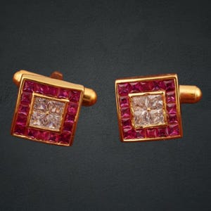 May include: A pair of gold-toned square cufflinks. Each cufflink features a central square of clear stones surrounded by a border of red stones. The cufflinks have a classic design and are set against a dark background.