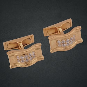 May include: A pair of gold-toned cufflinks with a rectangular design. Each cufflink features a curved central section adorned with small, clear gemstones. The cufflinks have a classic, elegant style, suitable for formal wear.