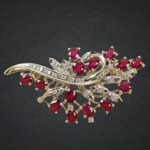 May include: A gold brooch with a floral design, set with oval-cut ruby gemstones and small diamonds. The brooch has a curved, flowing shape with intricate detailing. The background is dark.