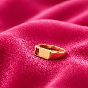 May include: A gold-toned ring featuring a smooth, polished band and a rectangular, deep red gemstone set horizontally into the flat top surface. The ring has a solid, substantial construction with a clean, minimalist design and a high-shine finish.
