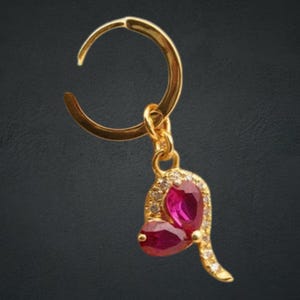 May include: A gold hoop earring with a pendant. The pendant has two oval, deep red gemstones and is embellished with small, clear gemstones. The earring is set against a dark background.