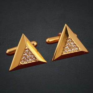 以下が含まれることがあります： 三角形のデザインの金色のカフスボタンのペア。各カフスボタンは、小さな透明な宝石のセクションを備えた三角形の形状を特徴としています。カフスボタンは光沢のある仕上がりで、シャツのカフに固定するためのポストが付いています。