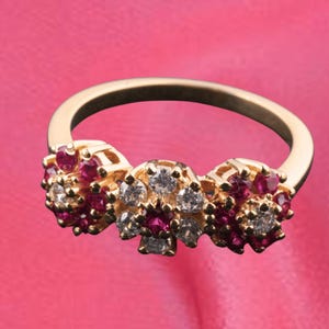 May include: A gold ring with three floral arrangements. Each arrangement is decorated with small, round, red gemstones and clear, round gemstones. The ring is set against a pink backdrop.