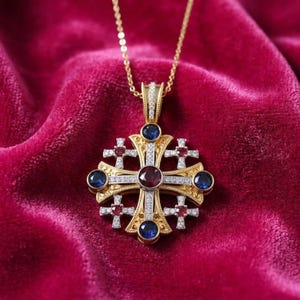 May include: A gold Jerusalem cross pendant necklace featuring a central round red gemstone, four smaller red gemstones on the cross arms, and four blue round gemstones at the tips, all accented with small white pavé-set stones on a gold chain.