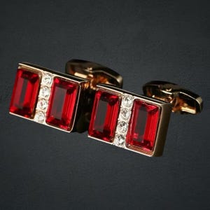 May include: Gold-coloured cufflinks featuring rectangular red gemstones and small clear crystals. These cufflinks have a classic design, suitable for formal occasions or adding a touch of elegance to any outfit.