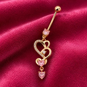 May include: A gold-toned belly button ring featuring a barbell with a small pink stone, a dangling heart-shaped charm encrusted with clear crystals, a central clear stone, and two additional pink heart-shaped stones hanging at the bottom.