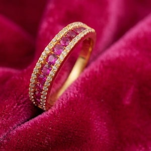 May include: A gold band ring featuring a central row of round, vibrant pinkish-red gemstones flanked by two delicate rows of tiny, clear white accent stones. The ring is crafted with a polished gold finish and a channel setting for the stones.