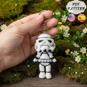 以下が含まれることがあります： 白と黒の編みぐるみストームトルーパーのキーホルダー。キーホルダーは、象徴的なヘルメットとアーマーの詳細なデザインが特徴です。「PDF PATTERN」というテキストが右上に見えます。キーホルダーは、緑の苔と小さな白と黄色の花の背景に対して保持されています。