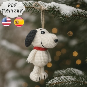 May include: A crocheted Snoopy Christmas decoration. The white Snoopy has black ears, a red collar, and a black nose. The ornament hangs from a twine string on a snow-covered evergreen branch. The image includes the text "PDF PATTERN".