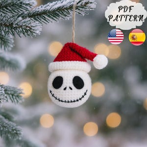 Puede incluir: Adorno navideño de Jack Skellington tejido a ganchillo. La esfera blanca tiene ojos negros y una sonrisa cosida. Está rematada con un gorro de Papá Noel rojo y blanco y un pompón blanco. El adorno cuelga de una cuerda de hilo natural. El texto "PDF PATTERN" está en el fondo.