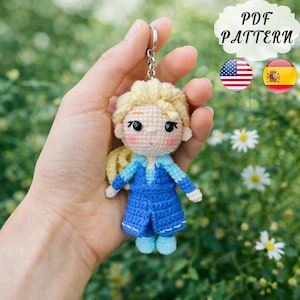 May include: A crocheted doll keychain with blonde hair and a blue dress. The doll is attached to a silver keychain. The image includes the text "PDF PATTERN" and flags of the United States and Spain.