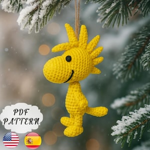 May include: A yellow crocheted Woodstock ornament with a black eye and a smiling mouth, hanging from a brown string. The ornament is suspended in front of a snowy evergreen branch. The text "PDF PATTERN" is visible.