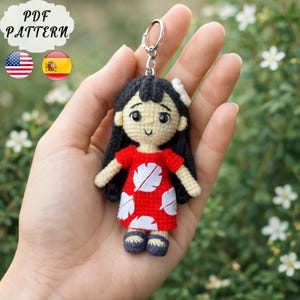 Könnte beinhalten: Gehäkelter Puppen-Schlüsselanhänger mit einer Figur in einem roten Kleid mit weißen Blattmustern. Die Puppe hat schwarzes Haar, eine weiße Blume und blaue Schuhe. Der Schlüsselanhänger hat einen silbernen Ring. Der Text "PDF PATTERN" und Flaggen sind ebenfalls vorhanden.