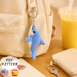 Puede incluir: Un llavero de tiburón tejido a ganchillo en azul y blanco, colgado de una mochila beige. El tiburón tiene boca rosa y ojos negros. En el fondo, una taza de plástico transparente con un batido amarillo. Se ve el texto "PDF PATTERN".