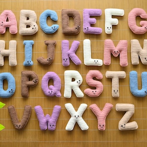 Peut inclure: Un alphabet complet de lettres crochetées de différentes couleurs, chacune avec un visage souriant. Les lettres sont disposées sur un tapis en bambou. L'image comprend une étiquette "PDF PATTERN". Les lettres sont dans des tons de rose, bleu, marron et blanc.