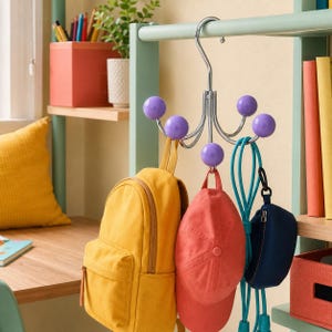 May include: A chrome-plated metal hook rack featuring four curved arms, each topped with a smooth purple ball. The rack is suspended from a horizontal bar and holds a yellow backpack, a coral baseball cap, a blue jump rope, and a small navy pouch.