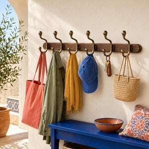 Puede incluir: Un perchero de pared con una tabla horizontal de madera oscura y seis ganchos de metal con acabado en latón antiguo. Los ganchos sostienen un bolso de tela color coral, una chaqueta verde oliva, una bufanda amarilla, una gorra azul, llaves y un bolso de paja.
