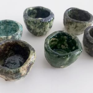 Op de afbeelding: Zes kleine, handgemaakte keramische kommen in tinten groen, blauw en grijs. Elke kom heeft een unieke, organische vorm en een gespikkelde glazuur, wat een rustieke en artistieke uitstraling creëert. De kommen zijn op een wit oppervlak geplaatst.