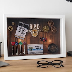 Puede incluir: Una vitrina enmarcada con objetos de temática policial, que incluyen una placa, monedas y llaves. La pantalla incluye una "WEAPONS LOCKER KEY CARD" y el texto "R.P.D. POLICE". También se ven una lámpara de escritorio negra, un portátil y gafas.