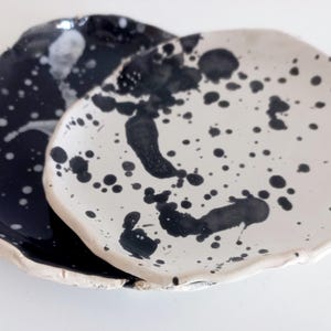 May include: Two ceramic plates, one black and one white, with irregular edges. Both plates are decorated with black paint splatters, creating a unique, artistic design. The plates are likely for serving food or as decorative pieces.