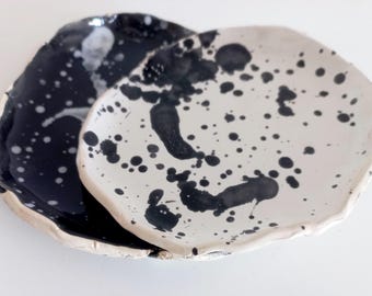 May include: Two ceramic plates, one black and one white, with irregular edges. Both plates are decorated with black paint splatters, creating a unique, artistic design. The plates are likely for serving food or as decorative pieces.
