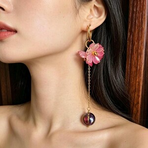 May include: A gold hoop earring featuring a translucent pink flower charm with a central gold bead. A delicate gold chain hangs from the flower, ending in a spherical, dark purple, iridescent bead that reflects light with a glossy finish.