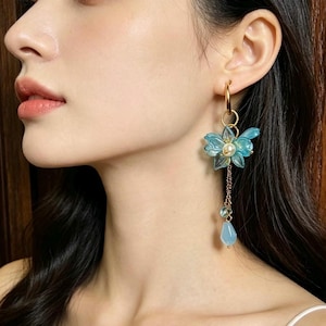 May include: A gold-toned hoop earring featuring a translucent blue flower charm with a central white pearl bead. A delicate gold chain hangs from the flower, ending in a small, light blue teardrop-shaped bead and a tiny matching blue accent.
