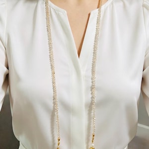 Könnte beinhalten: Eine lange Halskette mit zwei Strängen aus kleinen, hellen, facettierten Perlen, die von einem Choker-Band herabhängen. Der Choker ist mit einer einzelnen, unregelmäßigen weißen Perle besetzt, während die langen Stränge in goldfarbenen Akzenten enden.