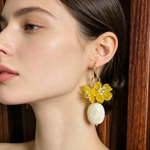 May include: A gold hoop earring featuring two translucent yellow flower charms with small white centres, suspended above a smooth, oval-shaped white stone bead. The earring is attached to a gold-toned metal hoop that secures to the earlobe.