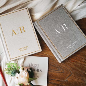 May include: Two personalized wedding vow books, one cream and one gray, with gold lettering and trim. The cream book has "AR" and "Alex's Vows 06.18.2025" printed on it. The gray book has "AR" and "Rachel's Vows 06.18.2025". A red pen and floral arrangement are also present.