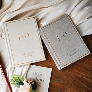 May include: Two hardcover vow books with fabric covers. The left book is cream with gold foil text reading J + O, OLIVIA'S VOW, and 04.13.2026. The right book is gray with black text reading J + O, JAMES'S VOW, and 04.13.2026. Both feature gold border lines.