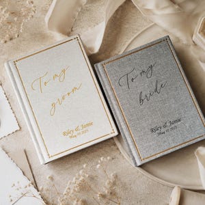 May include: Two rectangular hardcover books with fabric covers. The left book is cream with gold foil text reading To my groom, Riley & Jamie, May 10 2025. The right book is gray with black foil text reading To my bride, Riley & Jamie, May 10 2025.
