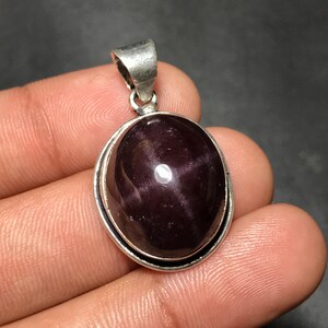 Pode incluir: Pingente de prata com uma pedra preciosa oval, roxa escura. A pedra preciosa tem um padrão em forma de estrela e é colocada em uma moldura de prata. O pingente tem uma argola de prata para uma corrente.
