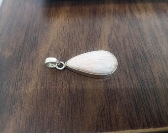 Könnte beinhalten: Ein tropfenförmiger Anhänger mit einem weißen, strukturierten Stein, der in einen silberfarbenen Metallrahmen gefasst ist. Der Anhänger hat oben eine Schlaufe für eine Kette. Der Schmuck wird auf einer dunklen Holzoberfläche präsentiert.