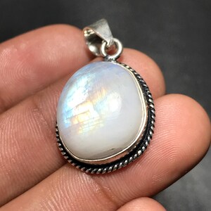 May include: A silver pendant with a large, oval moonstone. The moonstone has a pearly white color with iridescent flashes of blue and yellow. The pendant has a twisted rope design around the stone.