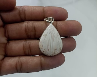 May include: A teardrop-shaped pendant with a silver bail. The pendant features a white stone with a unique, textured pattern and a silver border. The stone has subtle brown markings. The pendant is held in a hand.