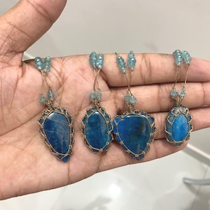 May include: Four blue gemstone pendants with gold-colored wire wrapping and light blue bead accents. The pendants are held in a hand, showcasing their unique shapes and textures. Each pendant has a different shape.