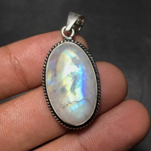 May include: Oval-shaped pendant with a large, iridescent moonstone. The stone displays a play of color with blues, greens, and yellows. The pendant has a silver-colored metal setting with a twisted rope design around the stone's edge.