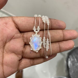 May include: A handcrafted macramé necklace with an oval moonstone pendant, displaying blue and white iridescence. The pendant is set within a white macramé design, complemented by white beaded details and fine white cords.