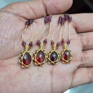 Può includere: Quattro collane fatte a mano con pietre preziose ovali rosso scuro incastonate in macramè color oro. Ogni collana ha un cordino dorato ed è adornata con piccole perline rosso scuro. Le collane sono esposte su una mano.