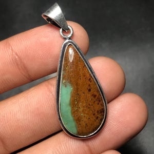 Puede incluir: Un colgante en forma de lágrima con una fianza plateada. El colgante presenta una piedra natural con una mezcla de colores marrón, verde y beige. La piedra está engastada en un marco de metal plateado.