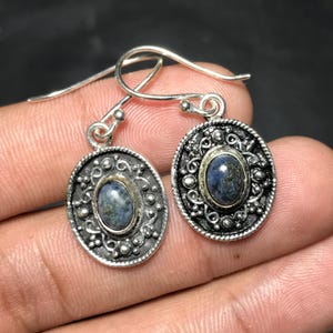 Op de afbeelding: Een paar zilverkleurige oorbellen met ovale, blauwe edelstenen. De oorbellen hebben een gedetailleerd, sierlijk ontwerp rond de edelstenen en een eenvoudige haaksluiting. De oorbellen worden tegen een donkere achtergrond gepresenteerd.