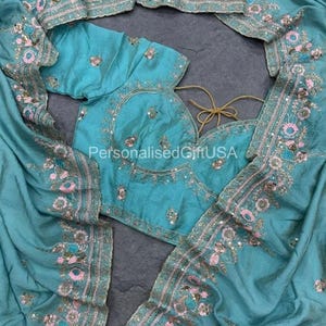 Op de afbeelding: Een blauwgroene sari-set bestaande uit een blouse en bijpassende doek met ingewikkeld bloemenborduurwerk in roze, blauwe en gouden draad. De stof is versierd met kleine reflecterende lovertjes. De tekst PersonalisedGiftUSA is zichtbaar.