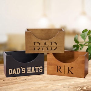Könnte beinhalten: Drei Holzkisten mit gebogenen Frontöffnungen. Eine Kiste ist dunkelbraun mit "DAD" und eingravierten Namen. Eine andere ist schwarz mit "DAD'S HATS" in Weiß. Die dritte ist hellbraun mit "RK" graviert. Alle Boxen stehen auf einer Holzoberfläche.