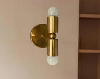 Puede incluir: Aplique de pared de latón con dos luminarias cilíndricas y dos bombillas globo blancas. El aplique tiene una placa trasera redonda y un diseño minimalista, ideal para interiores modernos.