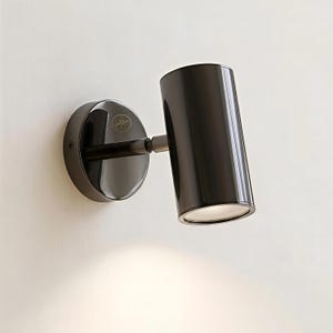 Puede incluir: Un foco de pared negro con una base circular y una lámpara cilíndrica. La lámpara está en ángulo y emite una luz brillante. La base tiene un pequeño emblema. El foco está montado en una pared blanca.