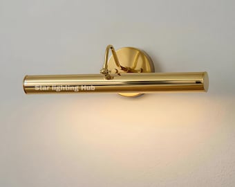 May include: A gold-colored picture light fixture with a cylindrical light bar and a circular wall mount. The fixture has a curved arm and a polished finish. The text "Star lighting Hub" is printed on the light bar.