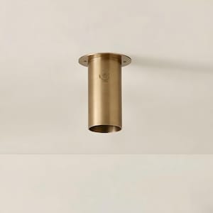 May include: A cylindrical, brass-colored ceiling light fixture. The fixture has a circular base with mounting holes and a cylindrical body. A small emblem is visible on the body. The light fixture is designed for recessed installation.