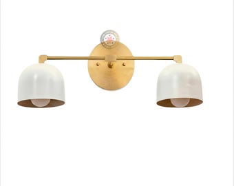 May include: A two-light wall sconce with a brass finish and white, bell-shaped shades. The fixture has a round backplate and a horizontal bar connecting the shades. The interior of the shades is gold, and each shade houses a light bulb.